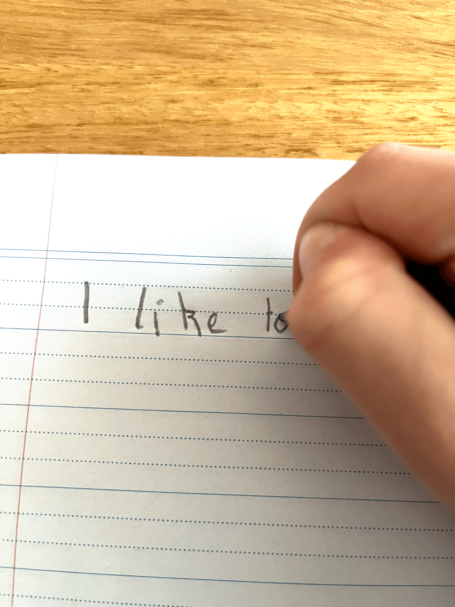 A child's hand writing the words 'I like to' in pencil on dotted-thirds handwriting paper